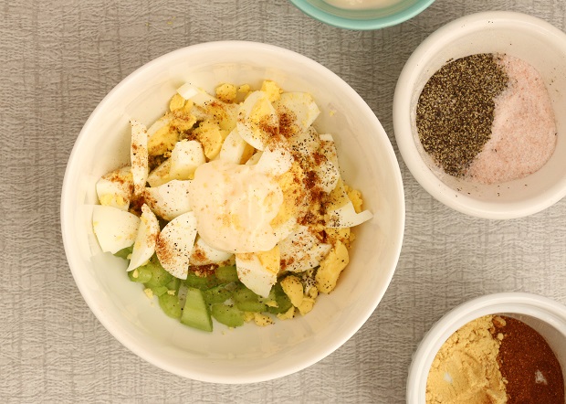 Celery and eggs in bowl topped with a dollop of mayo. Side bowls filled with paprika, dry mustard, salt, and pepper.
