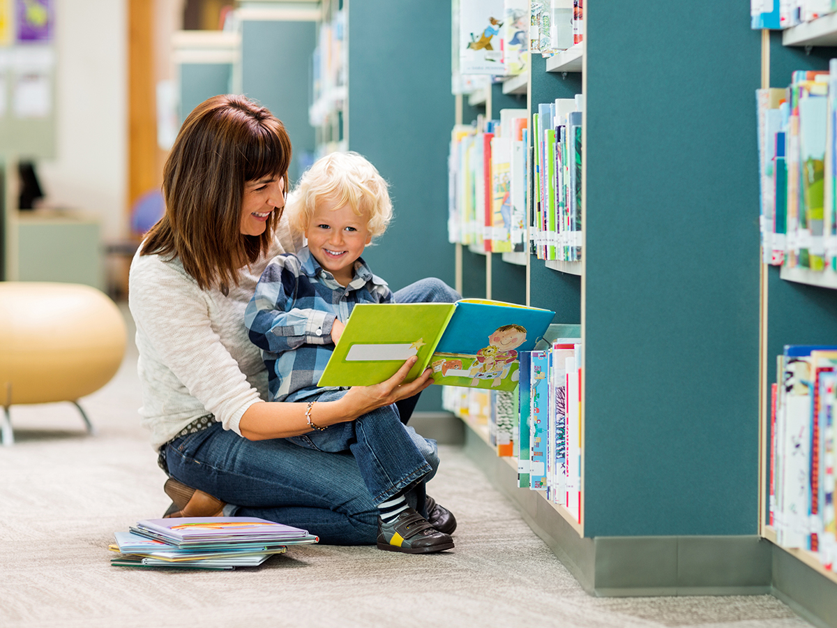 Smiling boy and teacher reading book by bookshelf in library