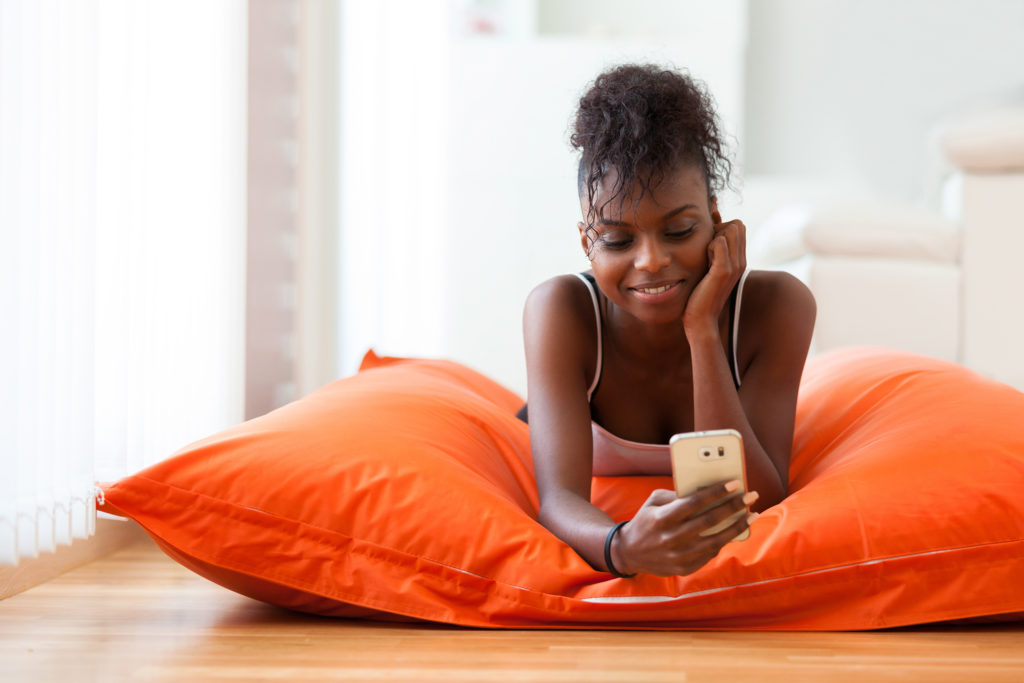African American woman laying on orange pillow while sending a text message on her smartphone
