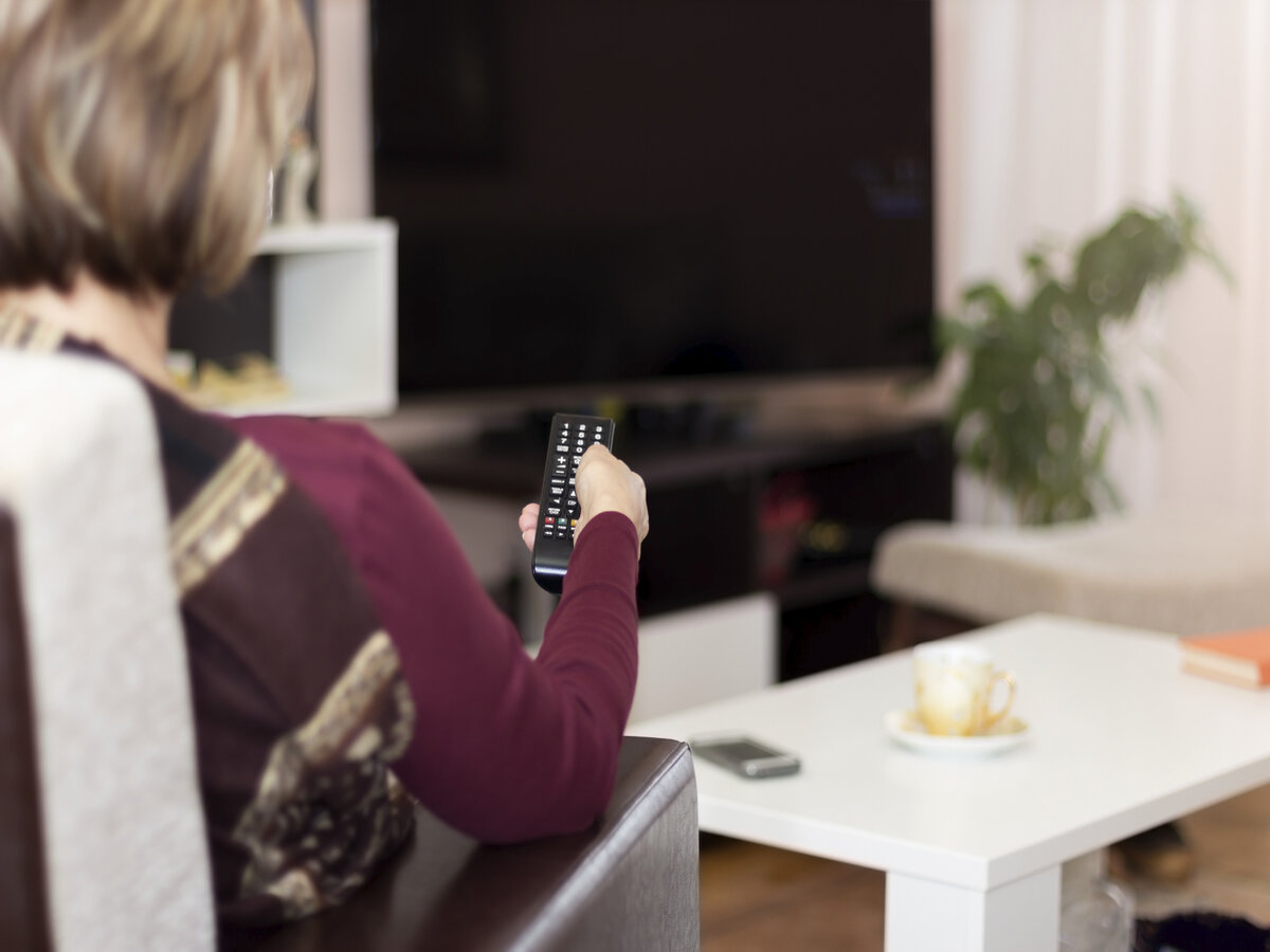 Woman on living room chair pointing remote control at a TV that won't turn on 