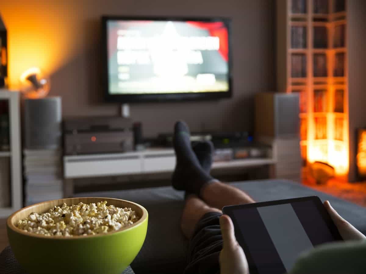 Person wearing black socks watching television while holding tablet with a green bowl of popcorn.