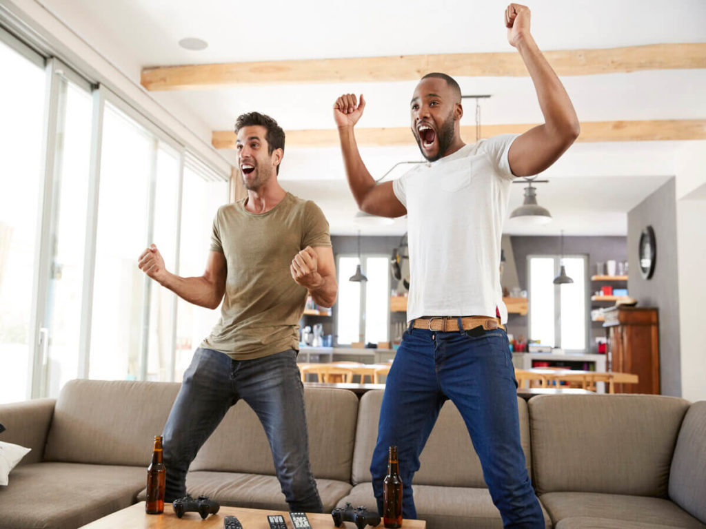 Two men are excited about a sports game, animated on their feet cheering in front of a taupe couch.