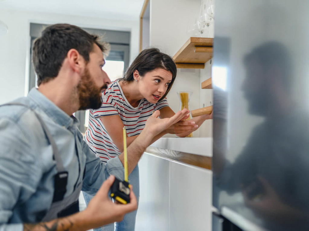 man with measuring tape and frustrated woman standing in front of fridge