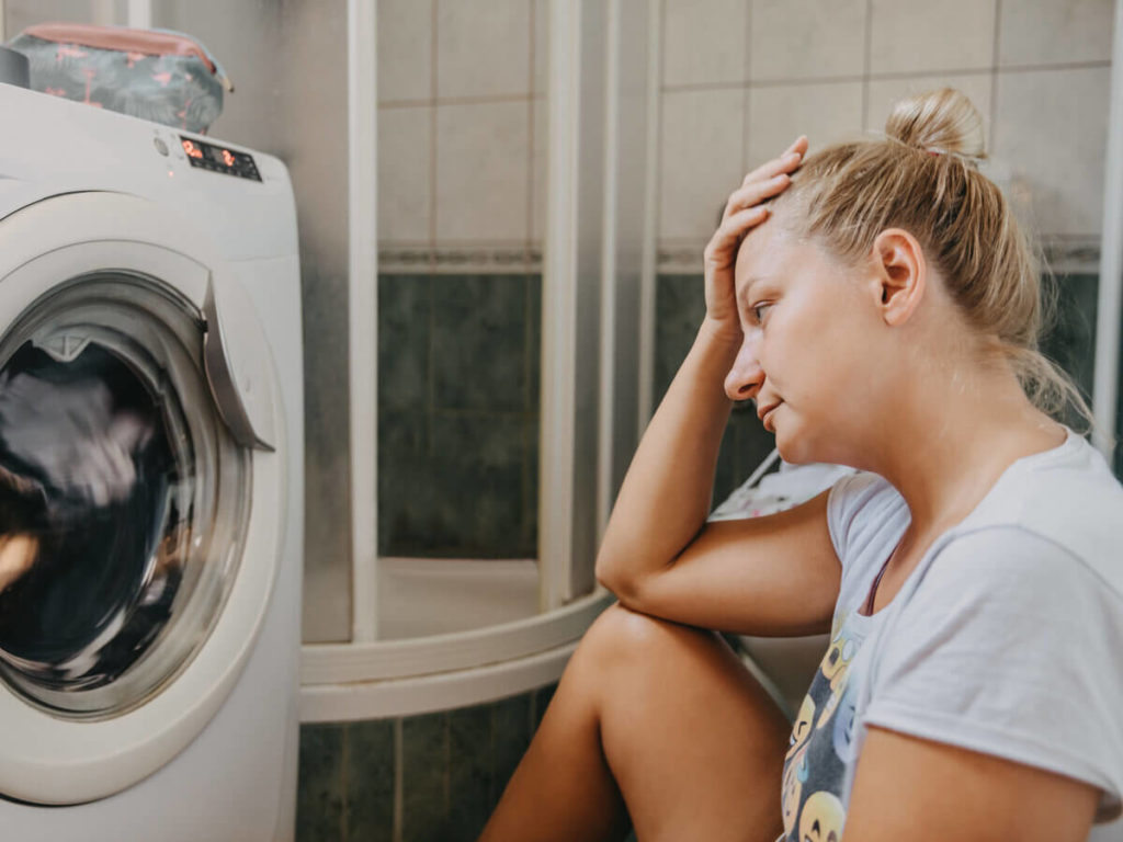 Woman sitting on laundry room floor with hand on head, looking upset at broken washing machine.