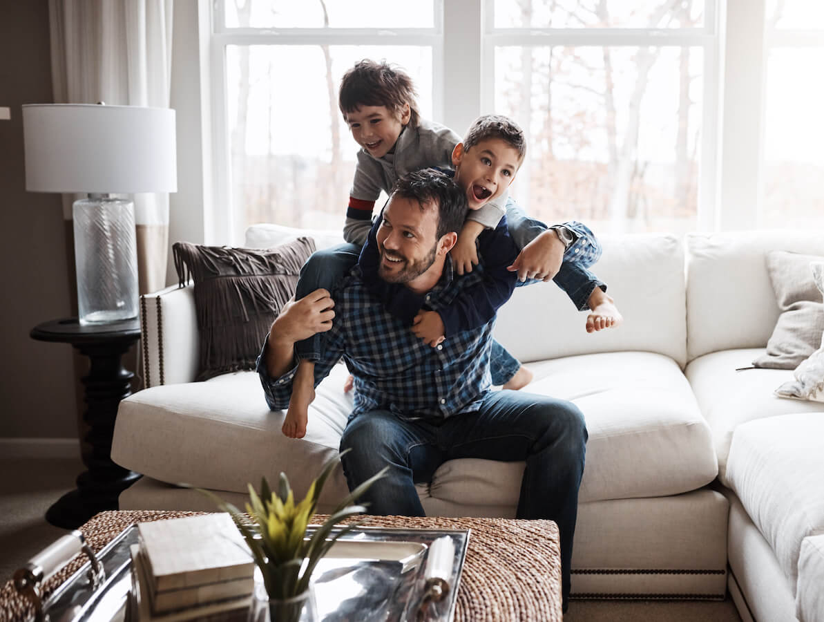 A man on a cream colored sectional plays with his two sons as they jump on his back.