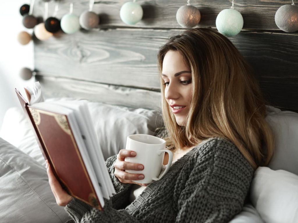 Woman sitting in bed with coffee mug reading a book.