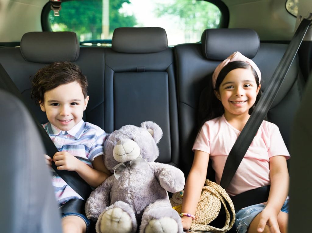 Small boy and girl sitting in backseat of car with teddy bear between them