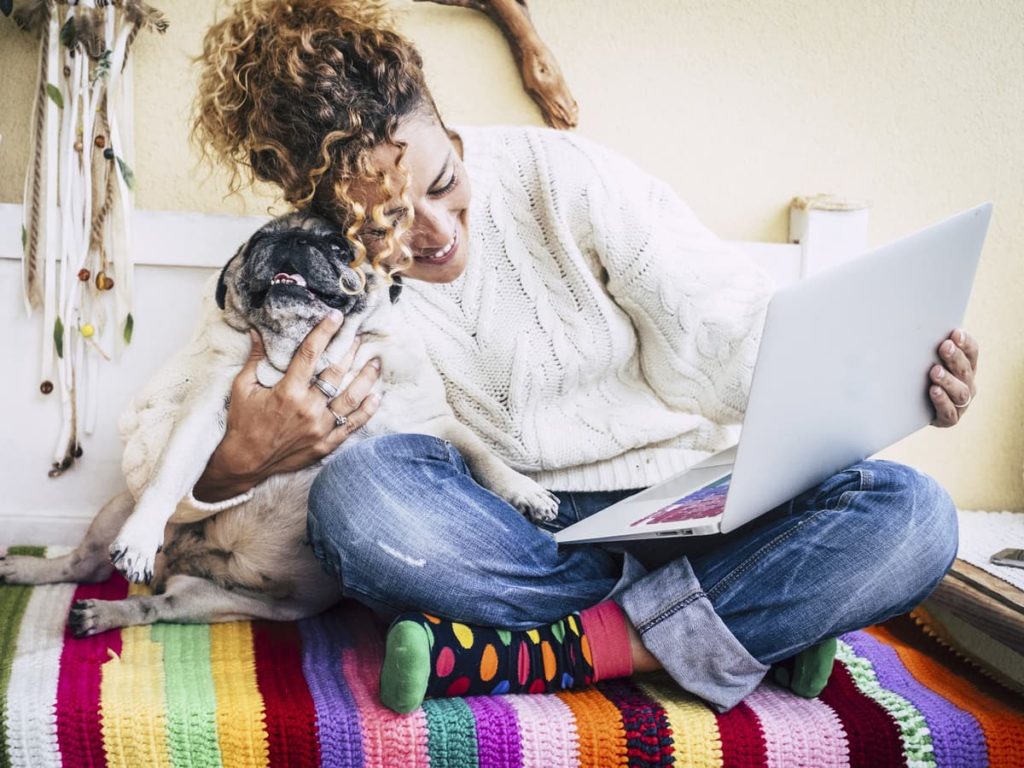 Woman on bed with dog while smiling at her laptop computer.