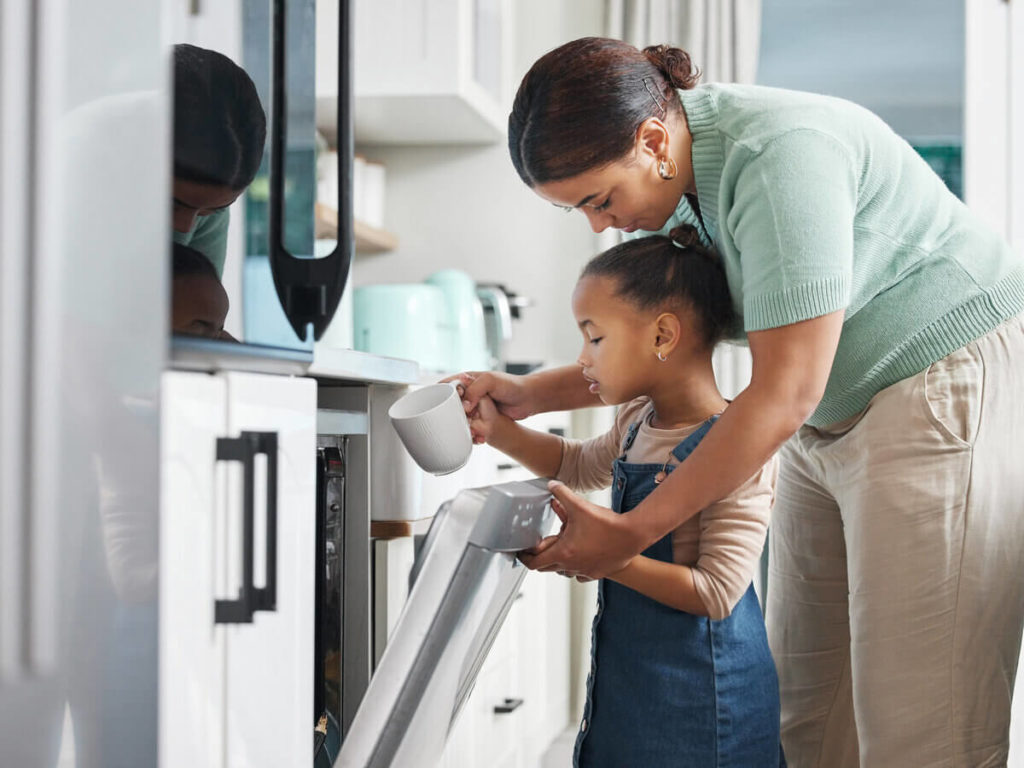 Mother stands behind young daughter as they load a dishwasher together.