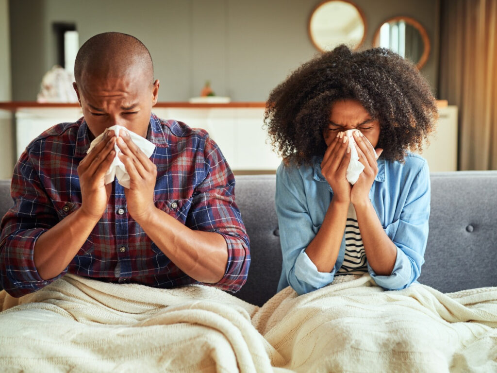 Allergy-ridden father and daughter sitting on couch while holding sneeze cloths