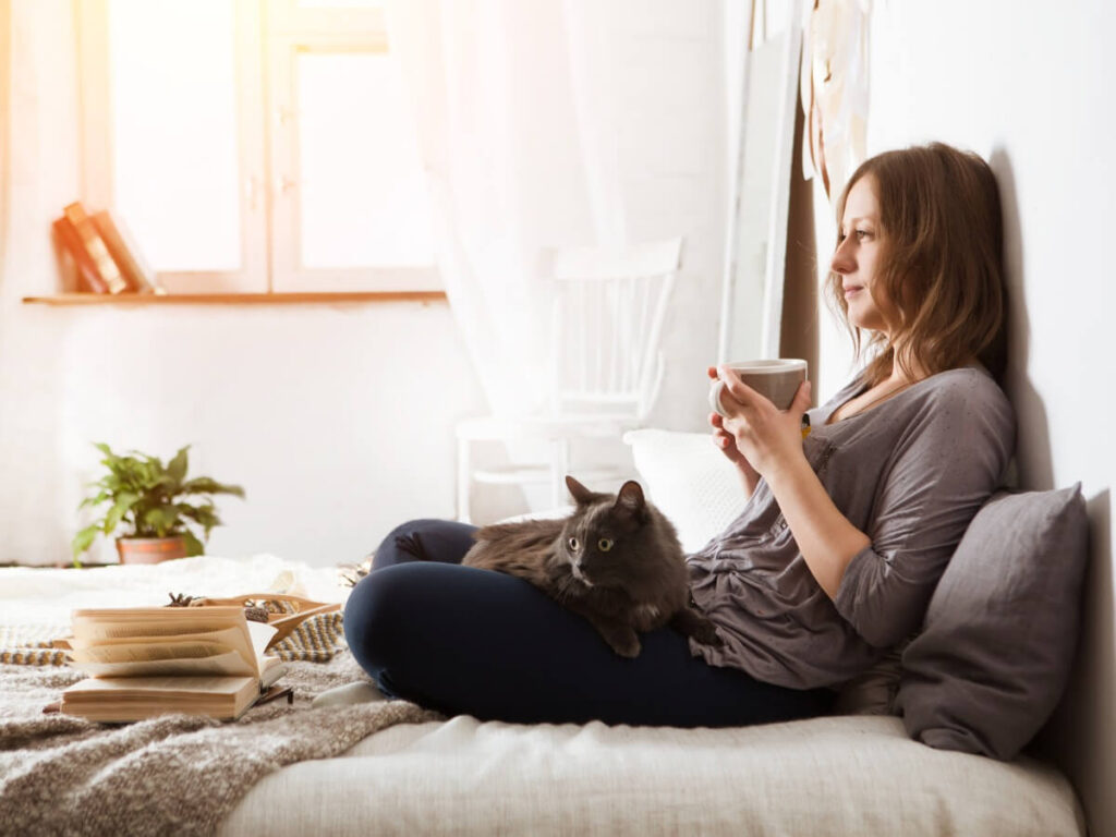 Woman sitting in bed in bedroom with cat on lap while holding a morning cup of coffee