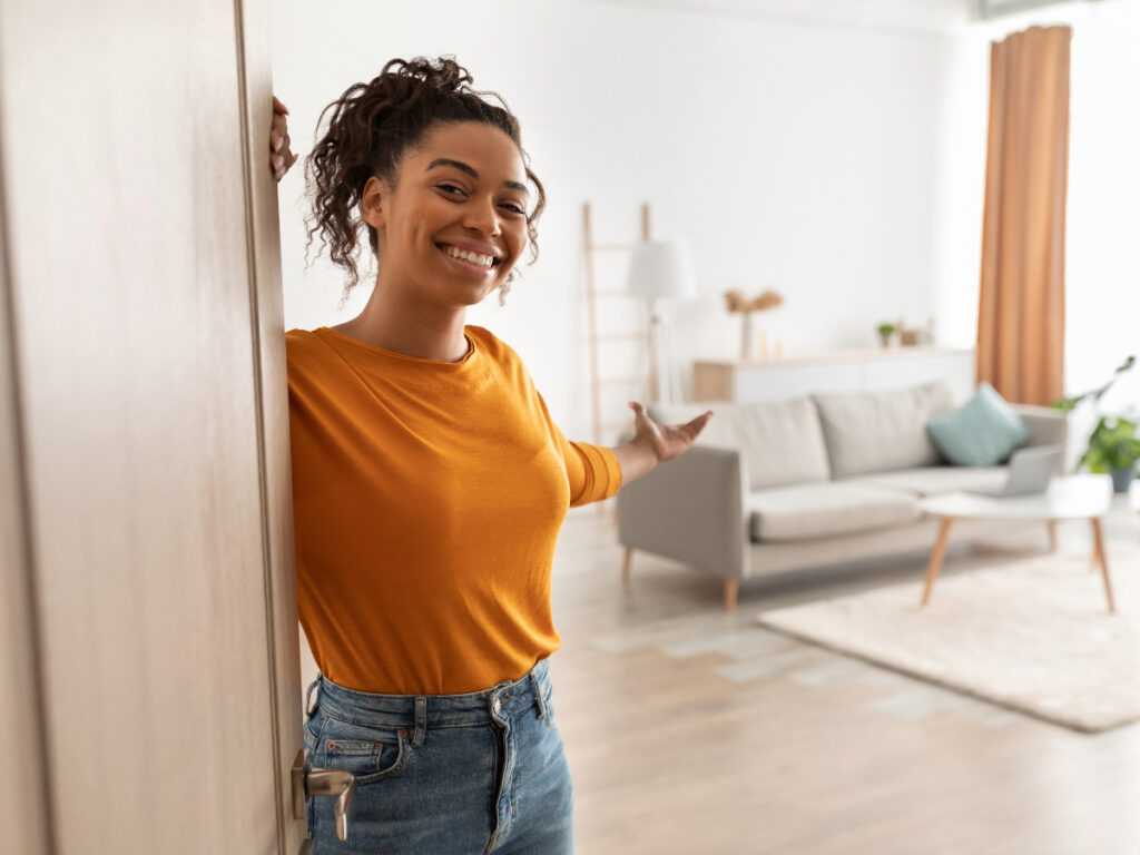 Smiling woman in orange top opening her apartment door, beckoning guests to come in