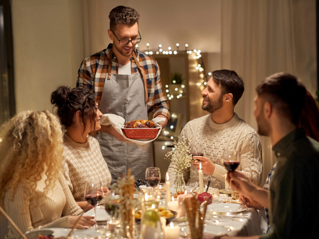 Group of smiling friends sitting around table for Thanksgiving with one young man standing with a freshly prepared Turkey