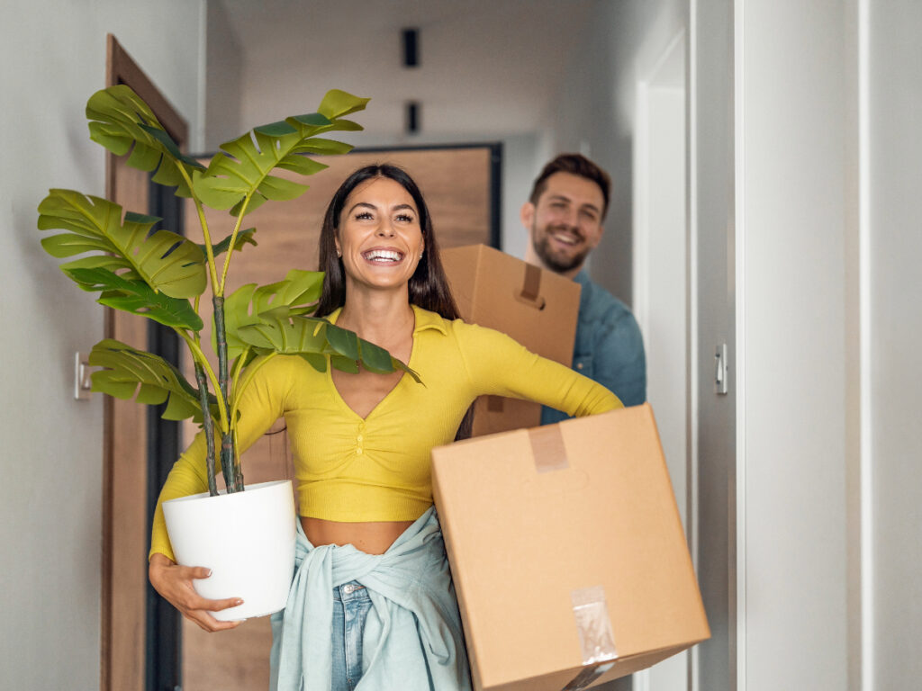 Smiling couple walking into a new living space. Woman is holding a box and mid-sized potted plant
