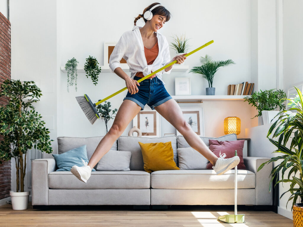 Cheerful woman with headphones and a broom (holding it like a guitar) captured jumping in mid air in her living room.