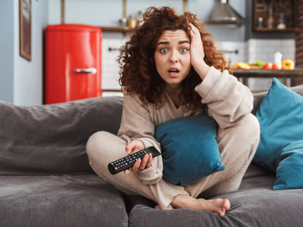Frustrated curly haired woman sitting on sofa with TV remote in hand.