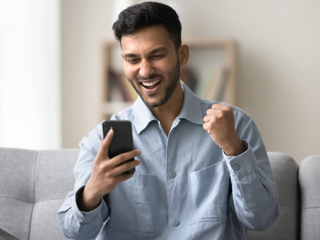Excited man sitting on sofa fist pumping while looking at his phone