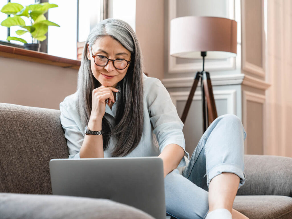 Woman with pleasant smile and glasses sitting on sofa in living room looking down at laptop.