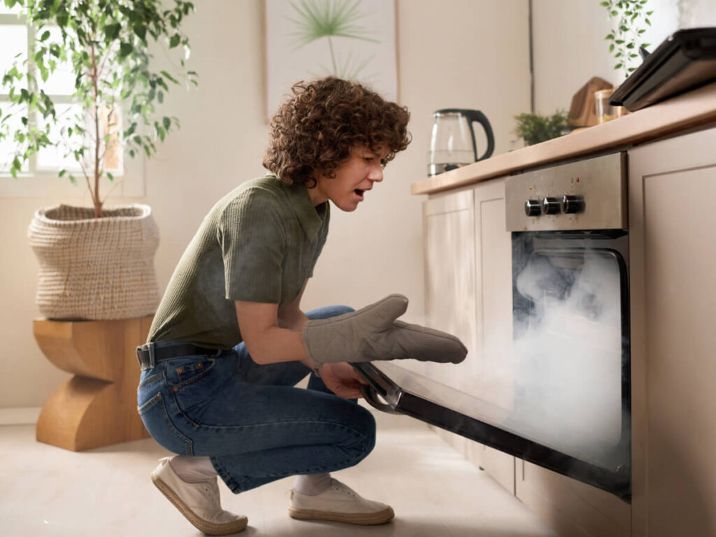 Confused woman squatting down with oven glove on in front of smoking oven