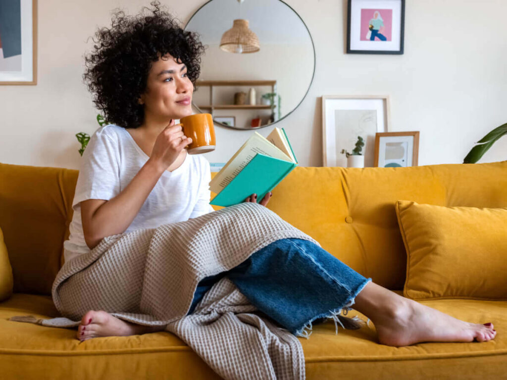 Woman sitting on stylish mustard yellow sofa with book and cup of coffee in hand while gazing out of a window