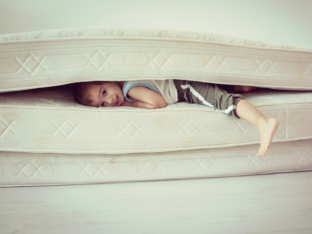 Toddler smiling at camera while sandwiched between a mattress and boxspring