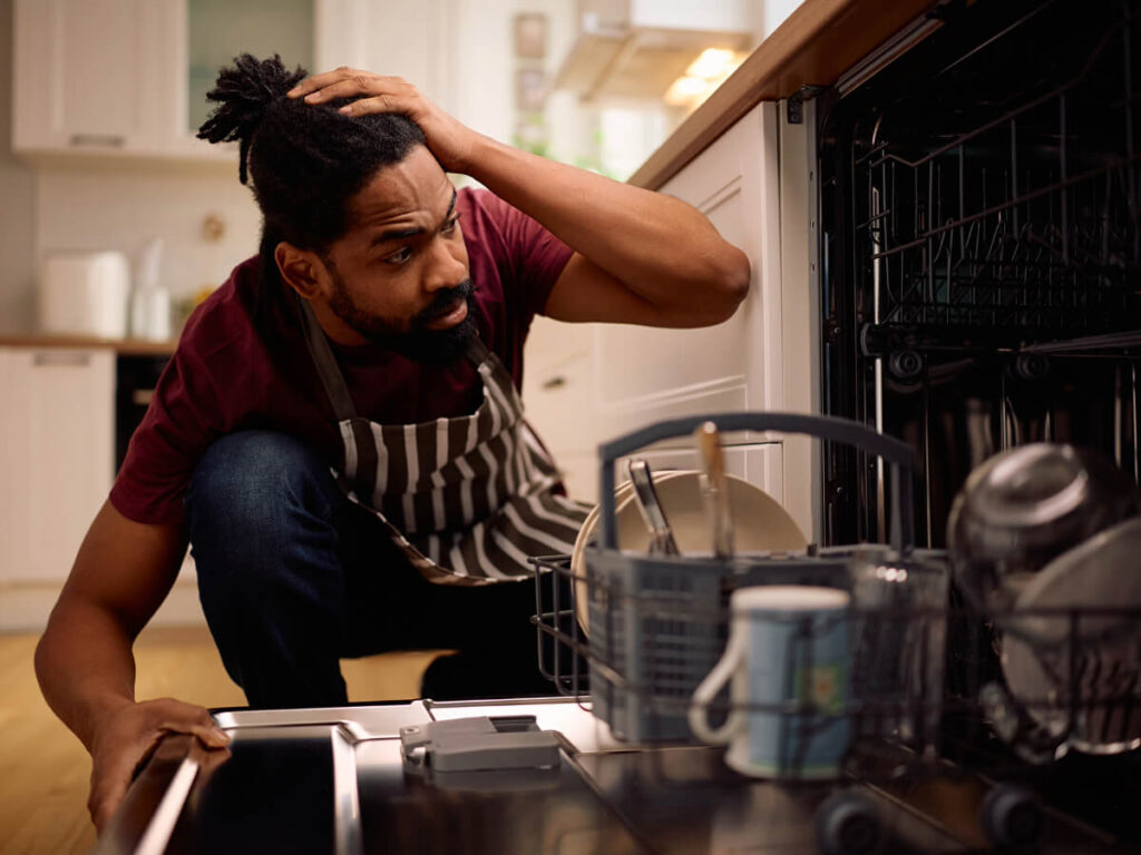 Perplexed man squatting and looking into dishwasher with this hand on his head as he looks inside of the appliance