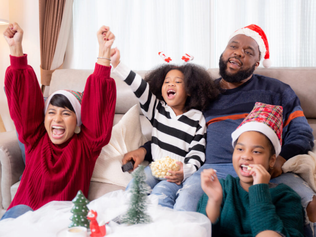 Two parents in living room with two children celebrating something they're watching on TV while wearing Santa hats