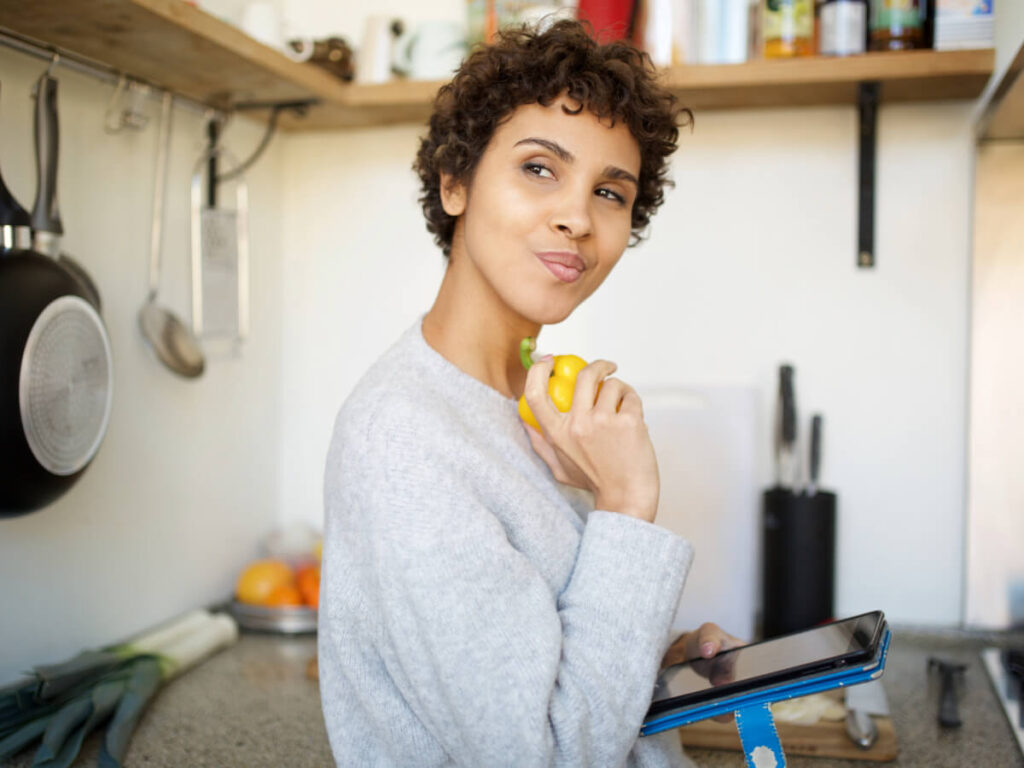 Woman contemplating something in her kitchen while holding a yellow bell pepper in one hand and an iPad in the other.