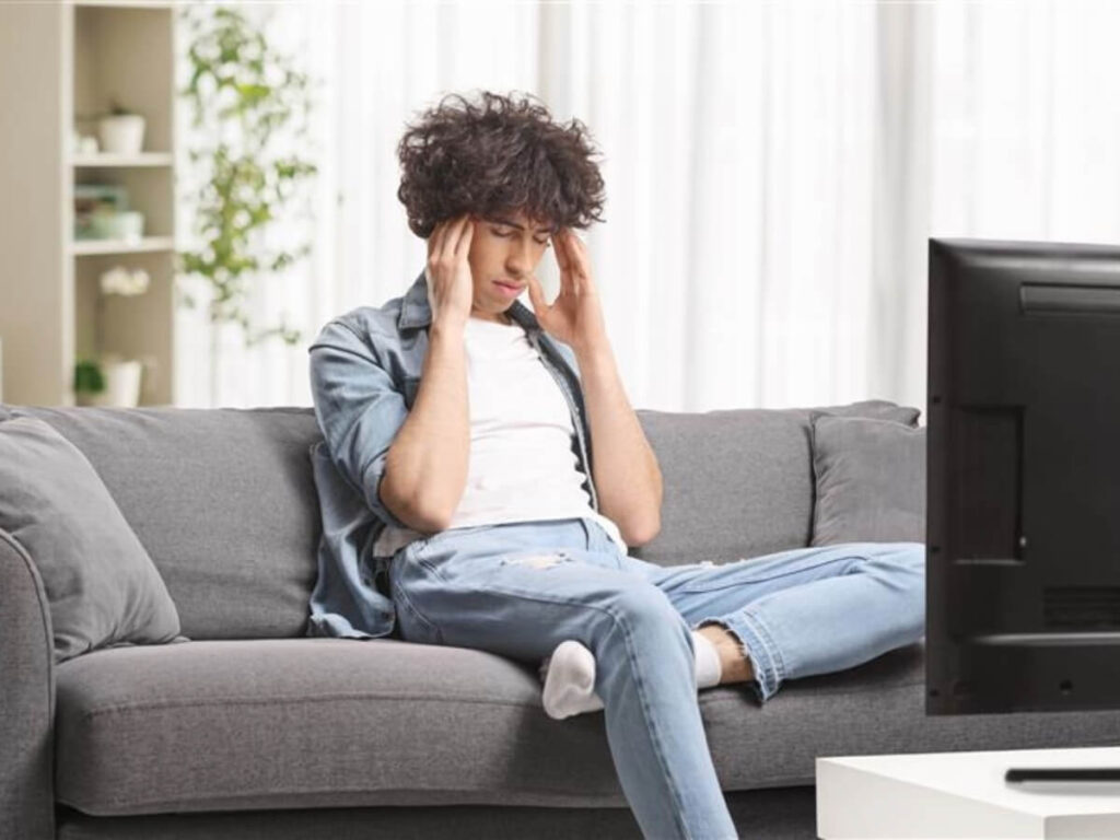 Man sitting on sofa with hands pressed against temples, frustrated while looking at TV