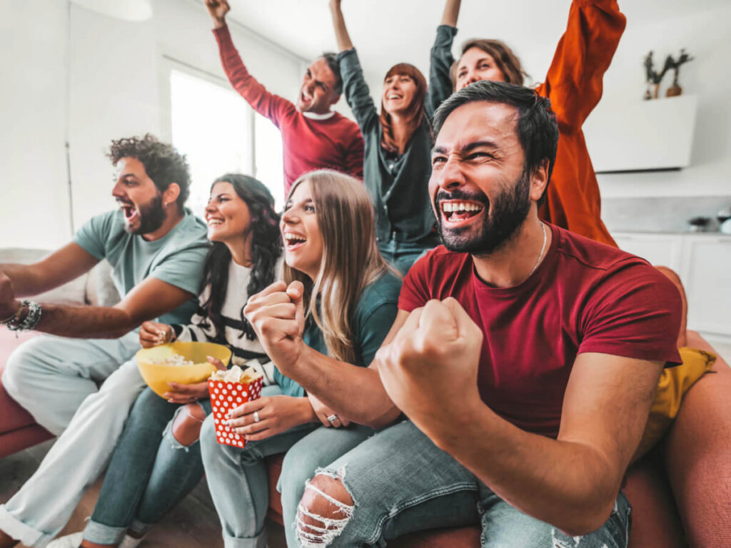 Group of millennial friends sitting on sofa cheering for a sports game while watching TV