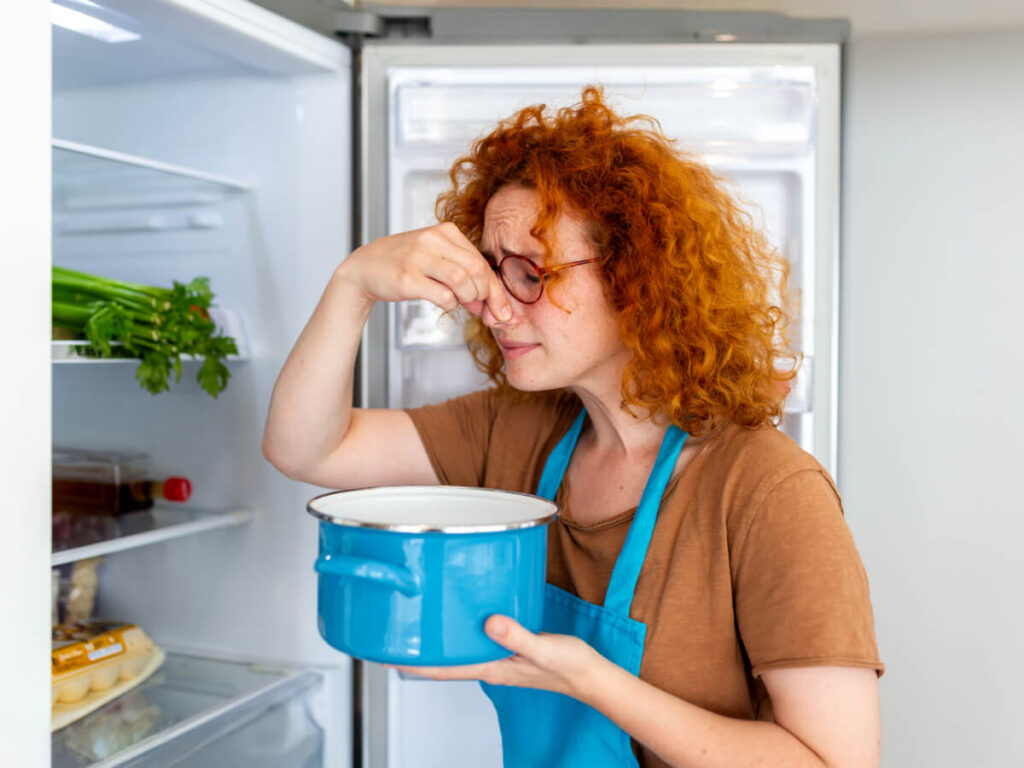 Woman standing in front of open fridge holding blue pot with one hand and her nose in the other