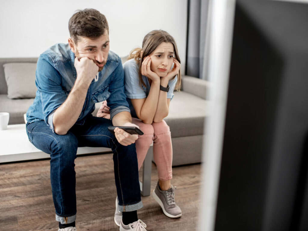 Young couple sitting on living room coffee table looking at TV with confusion and frustration