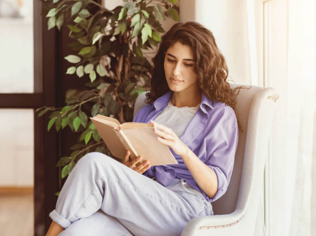 Woman relaxing in chair while reading a book in a sunny, cozy room
