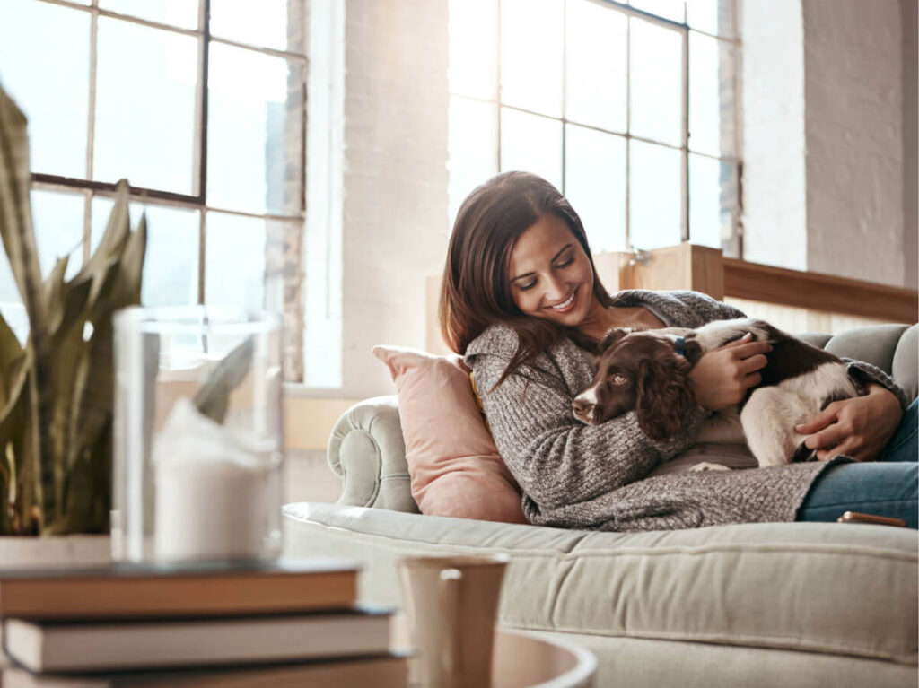 Woman on plush living room sofa with small puppy in her hand with large bay windows in the background