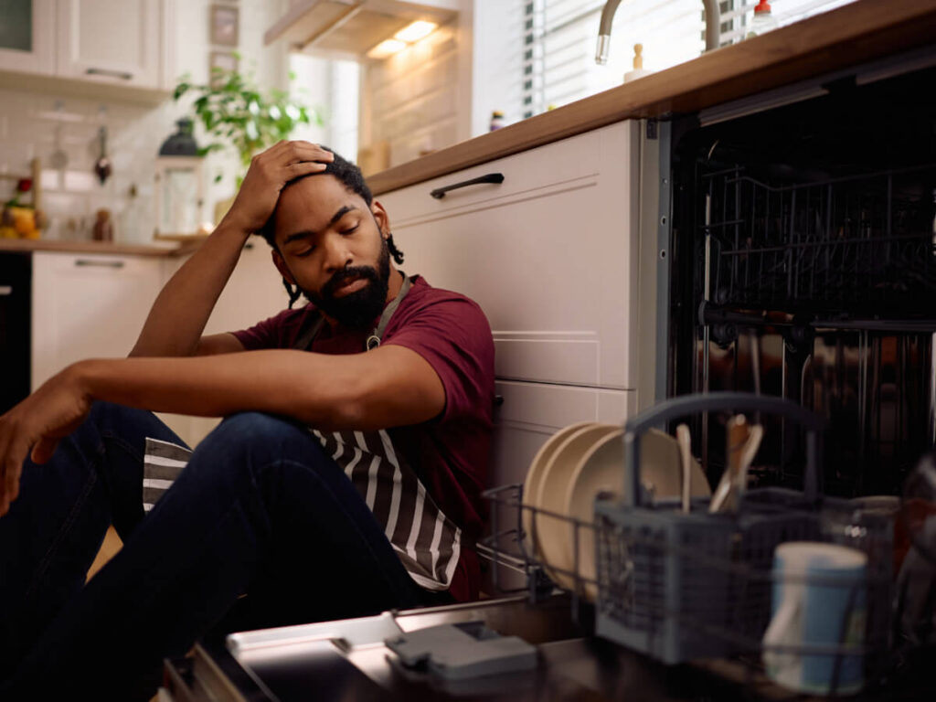 Frustrated man wearing apron with hand on head sitting next to open dishwasher