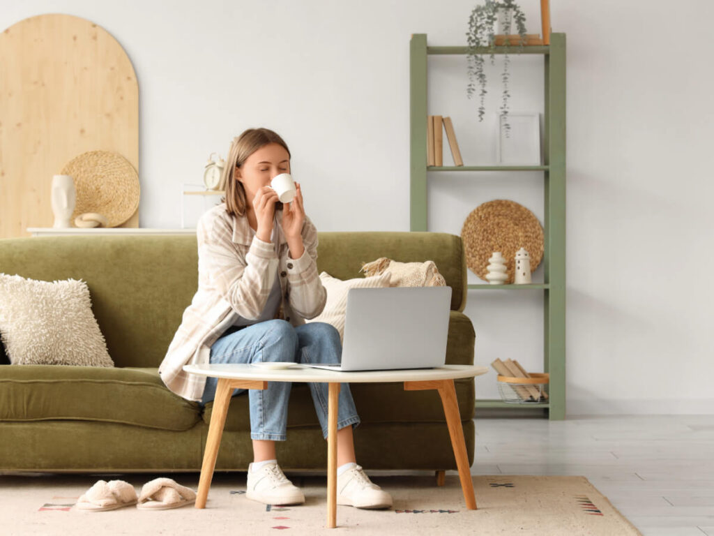 Young woman in minimalist living room sipping coffee on sofa with small coffee table holding laptop in front of her