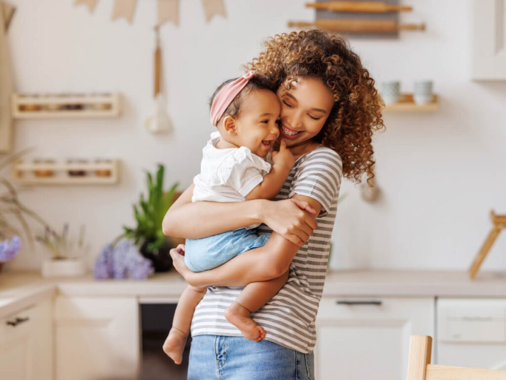 Smiling young woman with curly hair holding young child in arms in bright apartment kitchen