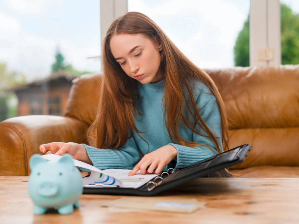 Young Woman Looking at Bills in Notepad with Piggy Bank on Table